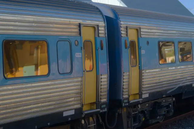 Older train model made of &ldquo;corrugated&rdquo; steel. A blue strip runs axially along the train, the doors are bright yellow and the windows are reflecting a yellow tint.