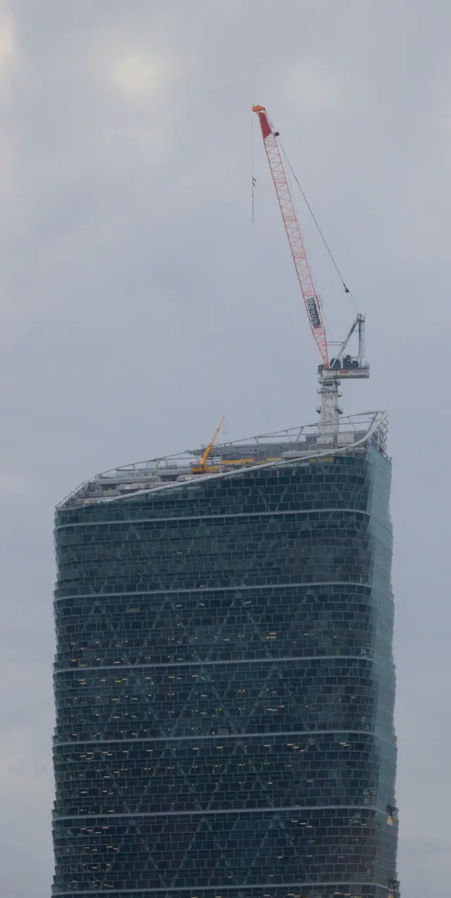 Top of a large glass building in construction. A small and a large lifting machine are visible atop it. Multiple floors can be seen, not just the top.